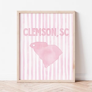 May include: A framed wall art print featuring a light pink and white vertical striped background. The text CLEMSON, SC is printed in a bold pink font above a watercolor-style pink silhouette of the state of South Carolina with a small white heart.