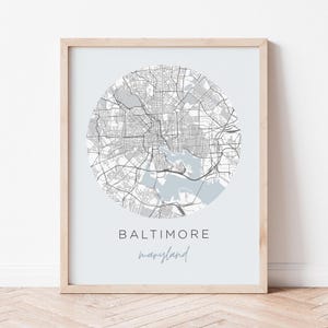 May include: A framed print showing a circular map of Baltimore, Maryland, in shades of grey with a pale blue water area. The city name is in a bold font, with "maryland" in a script font below.