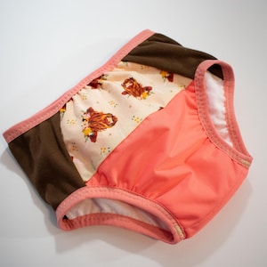May include: A pair of baby underwear with a patchwork design. The underwear features a brown panel, a peach panel, and a patterned panel with a floral design and a brown animal print. The edges are trimmed with a pink binding.