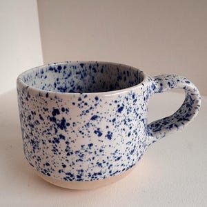 Könnte beinhalten: Eine Keramik-Tasse mit weißem Boden und einem gesprenkelten Muster aus dunkelblauen Punkten. Die Tasse hat eine abgerundete Form mit einem großen Henkel. Fassungsvermögen ca. 350 ml.