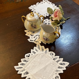 May include: A vintage tea set with a sugar bowl and creamer, featuring a floral design and gold accents. The set is placed on delicate white lace doilies, with a pink rose adding a touch of color. The items are arranged on a dark wooden surface.
