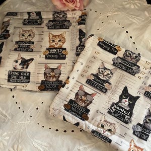 May include: Two white fabric squares with a cat mugshot design. Each cat has a "Wanted" sign with humorous crimes like "Cat Burglary" and "Wasting Lives." A pink rose rests on a white, eyelet-edged cloth.