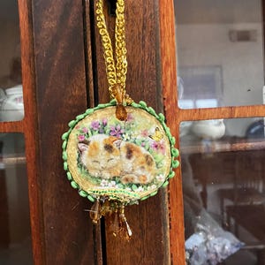 May include: A round, embroidered ornament featuring a sleeping calico cat surrounded by flowers and a butterfly. The ornament has a green and gold border, a gold chain, and dangling beads. It hangs from a black hook on a wooden door.