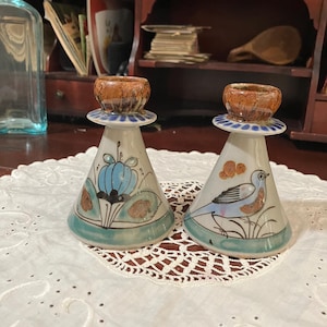 May include: Two conical ceramic candle holders with a cream-colored base. One features a blue floral design, while the other displays a blue bird. Both have a brown, textured rim at the top and a blue band around the base, finished with a glossy glaze.