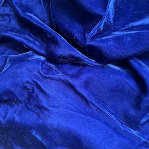 May include: A deep royal blue velvet fabric featuring a dense, soft pile with a distinct sheen. The material is draped in loose, flowing folds, creating varied light and shadow across the surface, highlighting the rich, saturated blue color.