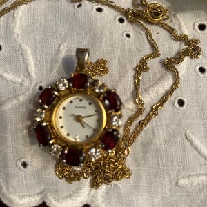 May include: A gold-tone pendant watch on a matching gold-tone chain. The watch face is white with the word QUARTZ printed on it. The bezel is decorated with alternating oval-shaped deep red stones and clear round stones in a circular arrangement.