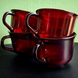 May include: Four ruby red glass mugs stacked on top of each other. Each mug has a rounded body, a curved handle, and a slightly flared rim. The mugs are made of a transparent material, allowing light to pass through.