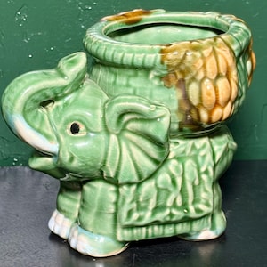May include: A green ceramic elephant planter with a textured surface. The elephant has a trunk raised and a basket-like container on its back. The planter has brown and tan accents and is suitable for small plants or decorative items.