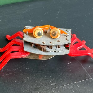 May include: A mechanical crab toy featuring a gray metal body with exposed internal gears, two orange plastic eyebrows, and two googly eyes. The toy has six bright red plastic legs extending from the sides, providing a stable base for the structure.