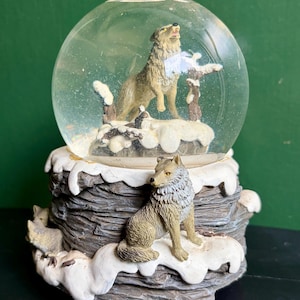 May include: A snow globe featuring a wolf howling inside a clear glass sphere. The base has a detailed, textured design with additional wolf figurines and faux snow. The wolves are shades of gray and brown, set against a snowy landscape.