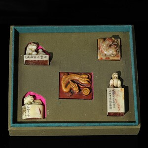 May include: A set of five carved stone seals arranged in a green tray. The collection includes two white stone seals with animal finials, one square seal with a carved dragon, one rectangular seal with a lion finial, and one cylindrical seal.