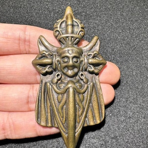 May include: A gold-colored metal pendant with a detailed design of a face with a sword and wings. The pendant is intricately carved and has a traditional style.