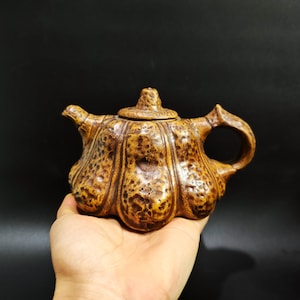 Puede incluir: Una tetera de cerámica marrón con una superficie texturizada y abultada, con forma de calabaza o calabaza. La tetera tiene una tapa y un asa.