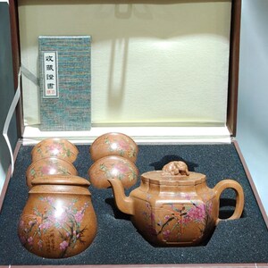 Puede incluir: Un juego de té de arcilla marrón que incluye una tetera con adorno en la tapa, un tarro con tapa y cuatro cuencos pequeños, todos decorados con ramas de flores rosas pintadas. El conjunto se presenta en una caja de color marrón.