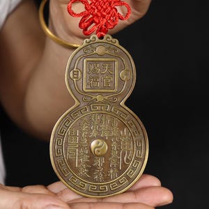 May include: A bronze-colored, double-sided pendant with Chinese characters and a yin-yang symbol. The pendant is suspended by a red knotted cord. The pendant is held in a person's hand.