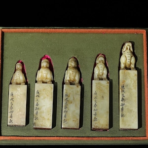 May include: A set of five rectangular off-white stone seals arranged in a green-lined box. Each seal features a carved animal figure on top and black Chinese characters inscribed vertically along the front face of the stone base.