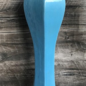 May include: A tall, light blue ceramic vase with a geometric shape. The vase has a wide base and a narrow neck.