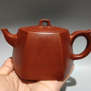 Könnte beinhalten: Eine rotbraune Keramik-Teekanne mit geometrischem Design. Die Teekanne hat einen gebogenen Ausguss, einen abgerundeten Griff und einen Deckel mit einer kleinen Schlaufe. Die Teekanne wird in einer Hand gehalten.
