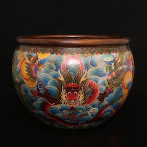 Peut inclure: Un bol cloisonné rond orné d'un motif vibrant représentant plusieurs dragons rouges, jaunes et verts sur un fond bleu de vagues stylisées. Le bol présente un bord en bois brun foncé et une bordure décorative sur le pourtour supérieur.