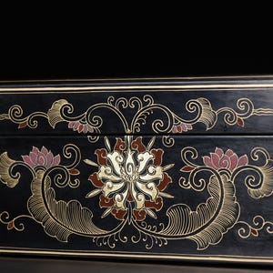 May include: A rectangular black lacquer box featuring intricate gold and muted red floral scrollwork patterns. The design includes symmetrical swirling lines and stylized lotus-like blossoms centered on the front panel with thin gold border lines.