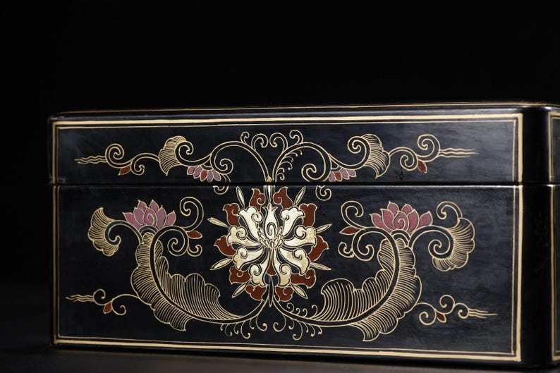 May include: A rectangular black lacquer box featuring intricate gold and muted red floral scrollwork patterns. The design includes symmetrical swirling lines and stylized lotus-like blossoms centered on the front panel with thin gold border lines.