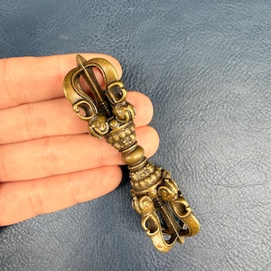 Puede incluir: Un objeto ritual vajra de metal color bronce con puntas simétricas intrincadas en ambos extremos, un agarre central con detalles de cuentas y un acabado de estilo antiguo desgastado con acentos de pátina oscura en toda la superficie.