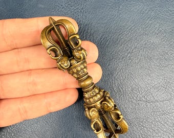 May include: A bronze-colored metal vajra ritual object featuring intricate symmetrical prongs at both ends, a central grip with beaded detailing, and a weathered, antique-style finish with dark patina accents throughout the textured surface.