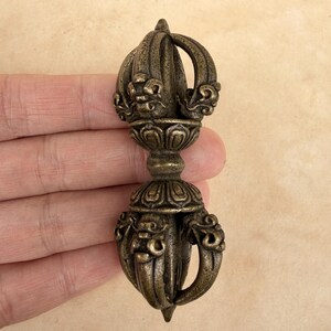 May include: A bronze-coloured vajra, a ritual object with ornate detailing. The double-ended sceptre features a central floral design and curved handles. The object is held in a hand against a neutral background, showcasing its intricate craftsmanship.