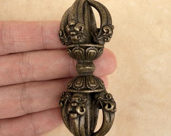 May include: A bronze-colored vajra, a ritual object with ornate detailing. The double-ended scepter features a central floral design and curved handles. The object is held in a hand against a neutral background, showcasing its intricate craftsmanship.
