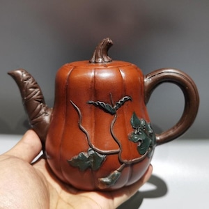 Puede incluir: Una tetera de cerámica marrón con forma de calabaza, con un pico y un asa curvados. La tetera está decorada con detalles de hojas y vides verdes. La tapa tiene un pomo en forma de tallo. La tetera se sostiene en una mano.