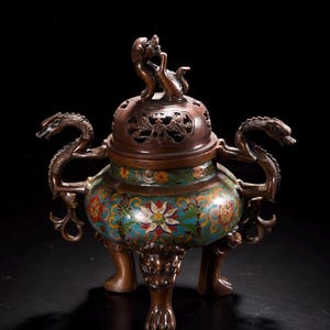 Op de afbeelding: Een antieke bronzen wierookbrander met een bloemen- en drakenontwerp. De brander heeft een gedetailleerd deksel met twee kleine figuren, drakenhandvatten en sierlijke poten. De body is versierd met kleurrijke emaille bloempatronen tegen een teal achtergrond.