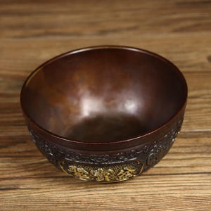 May include: A dark brown bowl with intricate carvings and gold accents. The bowl is made of metal and has a smooth, polished finish.