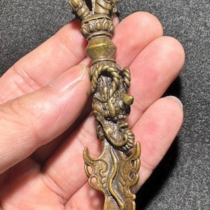 May include: A detailed bronze-colored ritual dagger, held in a hand. The dagger features intricate carvings, including a crown-like top, a dragon motif, and ornate blade details. The overall design suggests a ceremonial or decorative item.