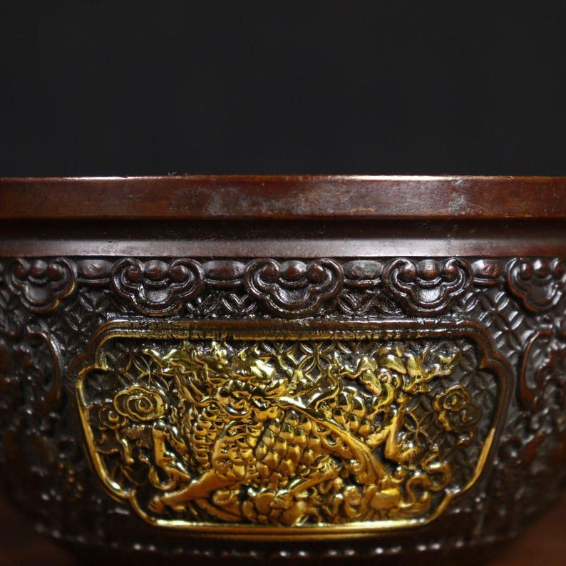May include: A round, dark brown bowl with intricate gold detailing. The bowl has a raised rim and a decorative design featuring a dragon.