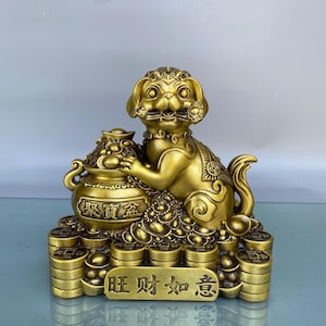 Puede incluir: Estatua dorada de un perro sosteniendo un pote del tesoro, sentado sobre pilas de monedas. La estatua tiene detalles intrincados y caracteres chinos. El diseño es ornamentado y simbólico, probablemente representando riqueza y prosperidad.