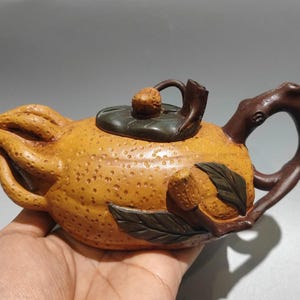 May include: A handcrafted teapot with a unique orange-like shape. The teapot is a warm yellow-orange color with a textured surface. It features a dark brown handle and lid, and is adorned with leaf and fruit details.