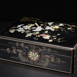 May include: A rectangular black lacquer box featuring an inlaid mother-of-pearl design of a bird perched on a vine with leaves on the lid. The sides are decorated with gold-colored painted floral motifs and swirling patterns, including Chinese calligraphy.