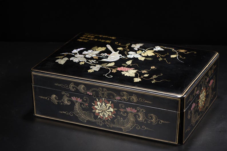 May include: A rectangular black lacquer box featuring an inlaid mother-of-pearl design of a bird perched on a vine with leaves on the lid. The sides are decorated with gold-colored painted floral motifs and swirling patterns, including Chinese calligraphy.