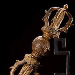 Op de afbeelding: Een bronzen vajra ritueel object met ingewikkeld metaalwerk, een centraal bolvormig gedeelte met gedetailleerde gravures en twee symmetrische uiteinden met tanden. Kleine turkooiskleurige inlays zijn rond de ringen verwerkt.
