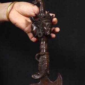 May include: A bronze-colored ritual dagger with intricate carvings. The handle features three faces and a dragon-like figure. The blade has a curved, crescent shape. The dagger is held against a dark background.