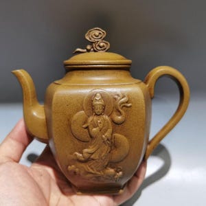 Könnte beinhalten: Braune Keramik-Teekanne mit detaillierter Reliefschnitzerei einer Figur auf der Vorderseite. Die Teekanne hat einen gebogenen Ausguss, einen Schlaufengriff und einen dekorativen Deckel mit Wirbeldesign. Die Teekanne wird in einer Hand gehalten.