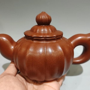 Op de afbeelding: Een roodbruine kleitheepot met een pompoenvormig lichaam met verticale ribbels. De theepot heeft een bijpassend deksel met een kleine, geribbelde knop, een gebogen tuit en een afgerond handvat aan de tegenoverliggende zijde.