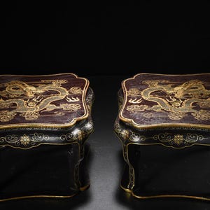 Peut inclure: Une paire de supports bas rectangulaires en bois présentant des surfaces sombres avec des illustrations complexes de dragons dorés sur les panneaux supérieurs. Les côtés sont finis en noir avec des détails floraux et des volutes dorées.