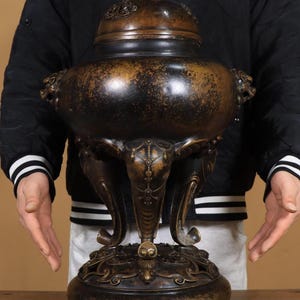Op de afbeelding: Een bronzen wierookbrander met een deksel, met ingewikkelde details en een donkere patina. De brander heeft een ronde basis, drie poten in de vorm van een olifant en handvatten in de vorm van een leeuwenkop. Bovenop het deksel staat een klein leeuwenbeeld. De totale hoogte is ongeveer 30 cm.