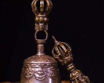 May include: A bronze bell and a dorje, a ritual object, are displayed against a dark background. The bell features intricate floral and geometric patterns, while the dorje has a detailed crown-like design. The objects are likely used in religious ceremonies.