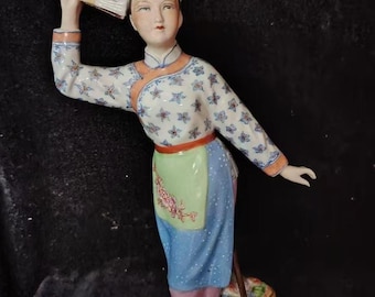 Op de afbeelding: Een porseleinen beeldje van een vrouw in traditionele Chinese kledij, met een waaier. Het beeldje draagt een top met bloemenpatroon, een blauwe rok en een roze broek. Een mand wordt in haar andere hand gehouden. Het beeldje staat op een witte voet met gouden rand.