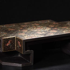 Peut inclure: Une table basse rectangulaire avec une finition noire et des motifs floraux dorés et rouges complexes sur le dessus. La table présente un design de base géométrique et étagé avec des motifs décoratifs assortis sur les panneaux latéraux.