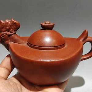 May include: A reddish-brown ceramic teapot with a rooster-shaped spout and handle. The lid is round with a small knob. The teapot is held in a hand, showcasing its detailed craftsmanship and unique design. This decorative teapot is ideal for tea enthusiasts.