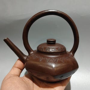 May include: A brown ceramic teapot with a large, arched handle and a spout. The lid is decorated with geometric patterns. The teapot is held in a hand, and the base has Chinese characters.