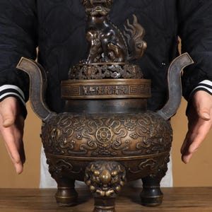 Op de afbeelding: Een bronzen wierookbrander met een gedetailleerd ontwerp. De brander heeft een zittend mythisch wezen bovenop, ingewikkelde patronen en handvatten. Het stuk wordt ondersteund door drie sierlijke poten en wordt waarschijnlijk gebruikt voor religieuze of decoratieve doeleinden.